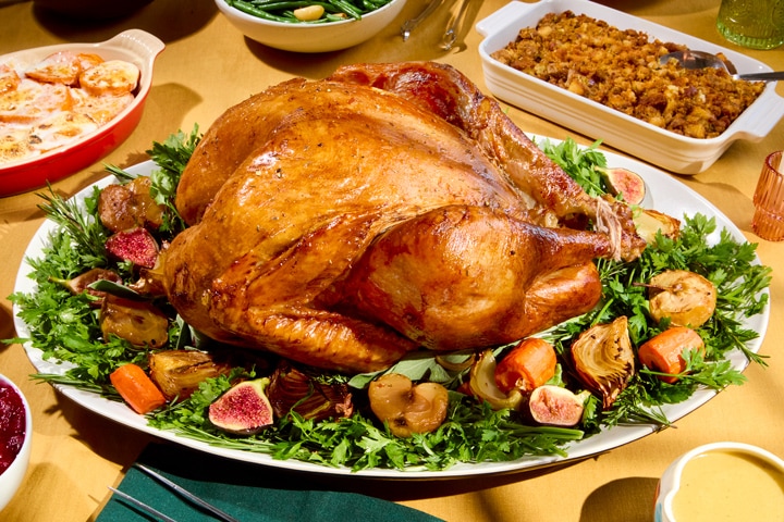 Whole roasted turkey on a platter with greens, roasted vegetables, and fresh figs